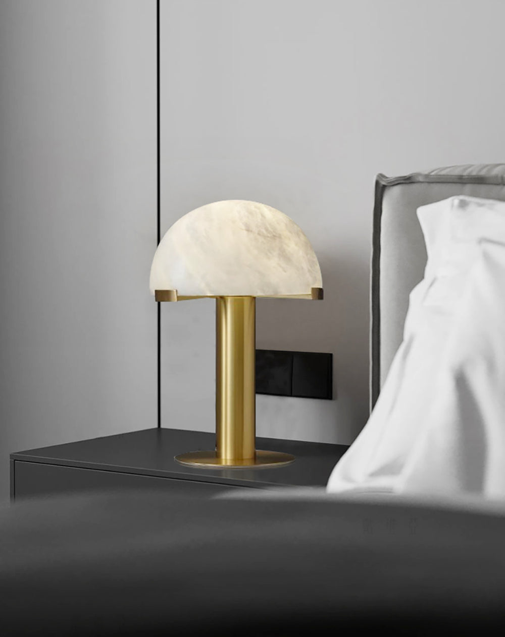 Gold Clérachic table lamp on a dark bedside ledge, with a grey upholstered headboard and crumpled white bedding behind.