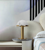 Wider view of the gold Clérachic table lamp on a round marble side table, with a cream headboard and striped bedding.
