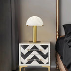 Gold Clérachic table lamp lit on a white nightstand with black chevron drawers, beside a dark bed and curtain.
