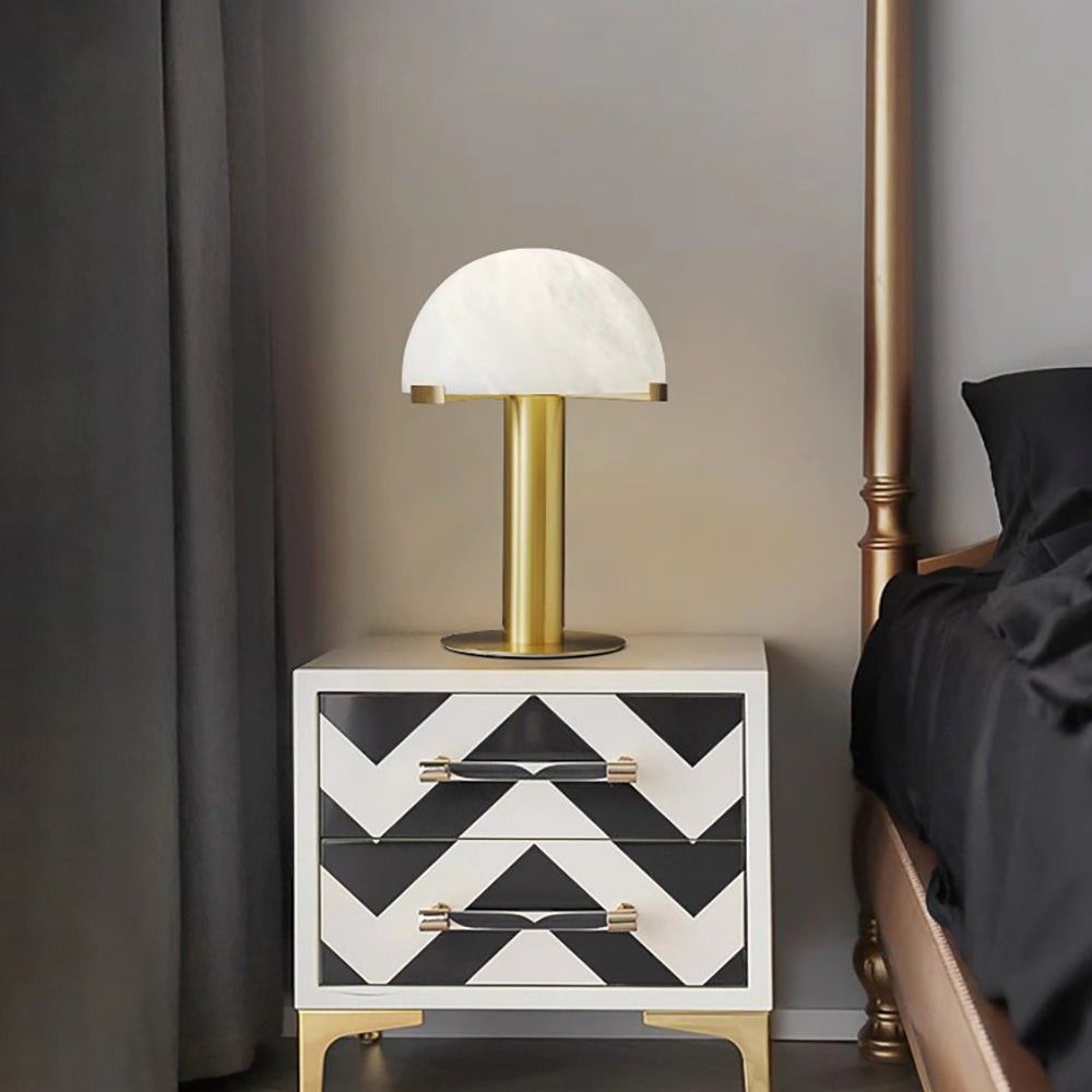 Gold Clérachic table lamp lit on a white nightstand with black chevron drawers, beside a dark bed and curtain.