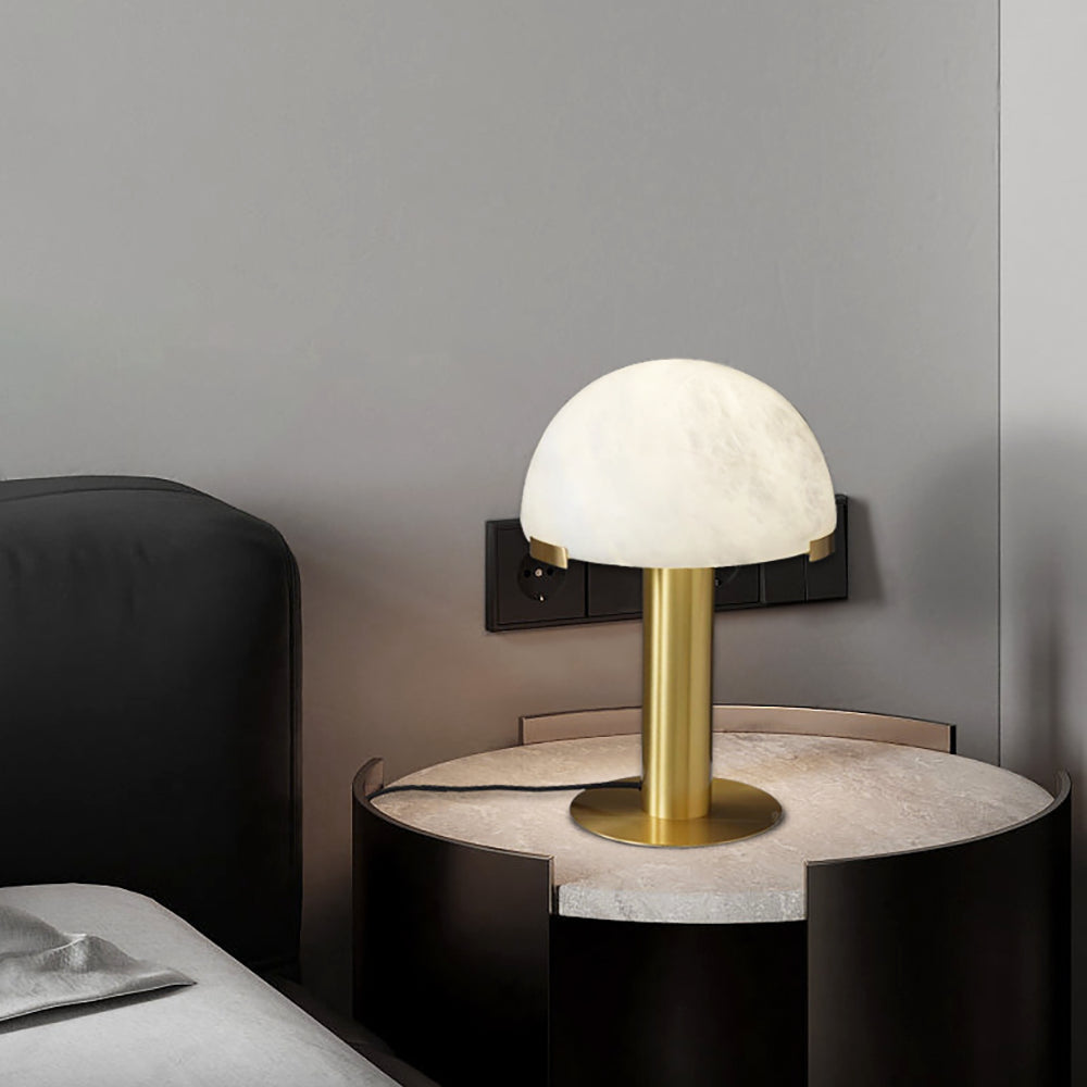 Gold Clérachic table lamp on a round pale-topped bedside table, with a wall socket and a dark headboard behind.