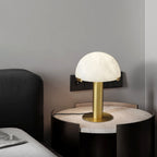 Gold Clérachic table lamp on a round pale-topped bedside table, with a wall socket and a dark headboard behind.