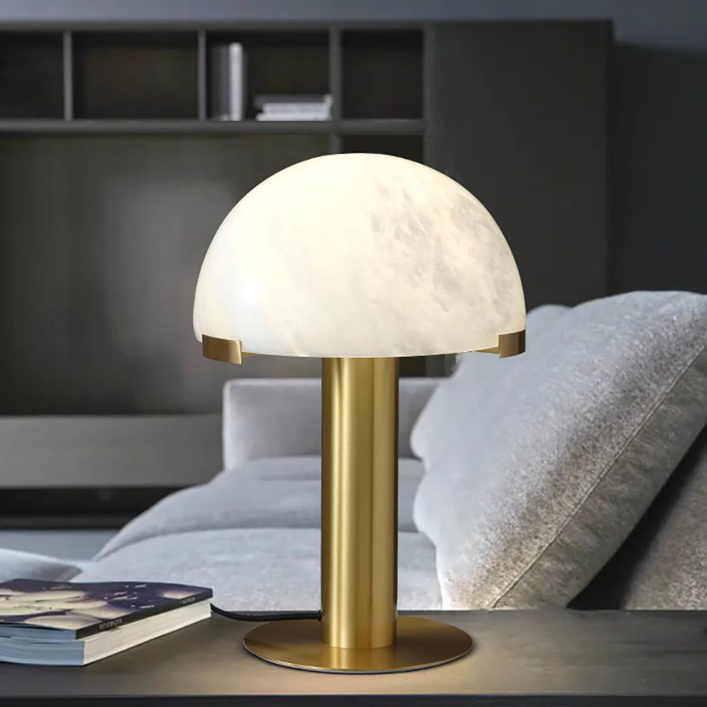 Close view of a gold Clérachic table lamp on a dark table, with a grey sofa cushion and an open magazine at the left.