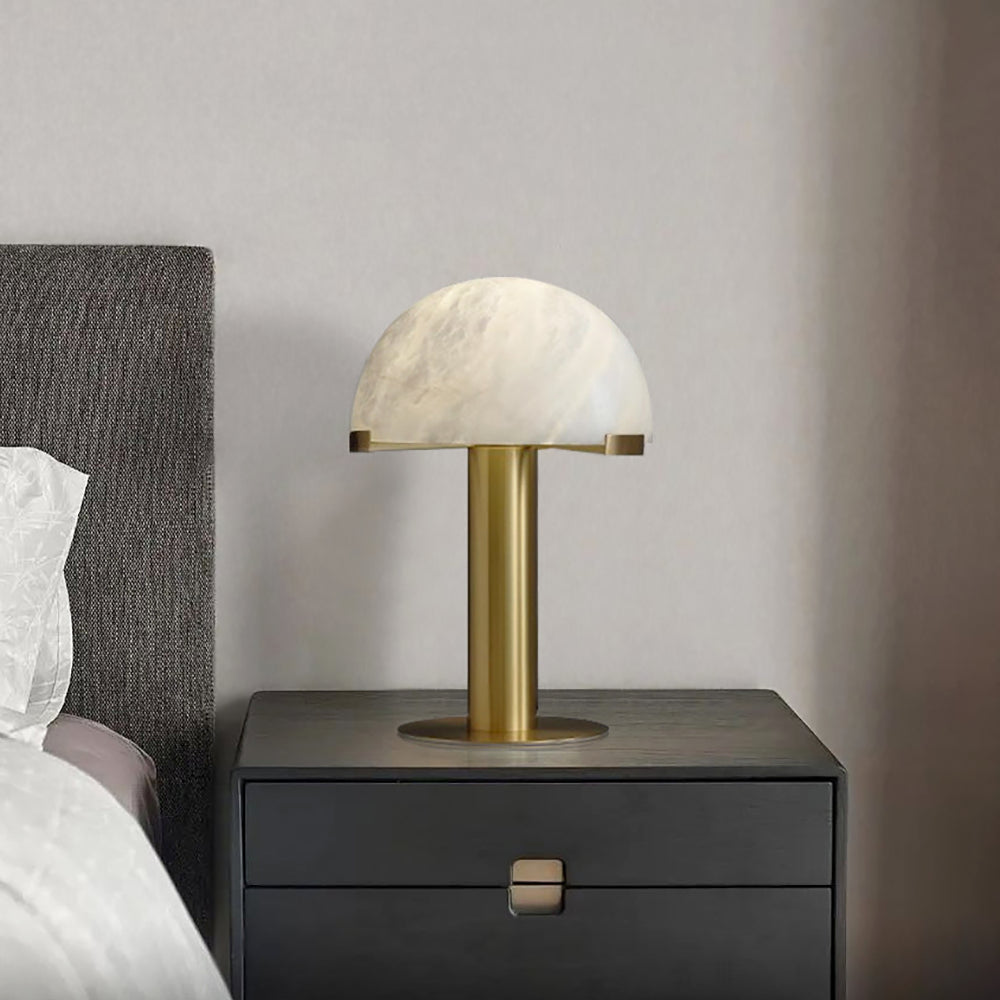 Gold Clérachic table lamp on a black two-drawer nightstand, against a beige wall with a grey headboard to the left.