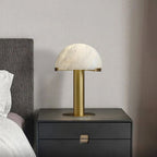 Gold Clérachic table lamp on a black two-drawer nightstand, against a beige wall with a grey headboard to the left.