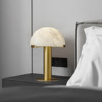 Gold Clérachic table lamp on a dark bedside surface, a black wall switch plate behind it and white pillows to the right.