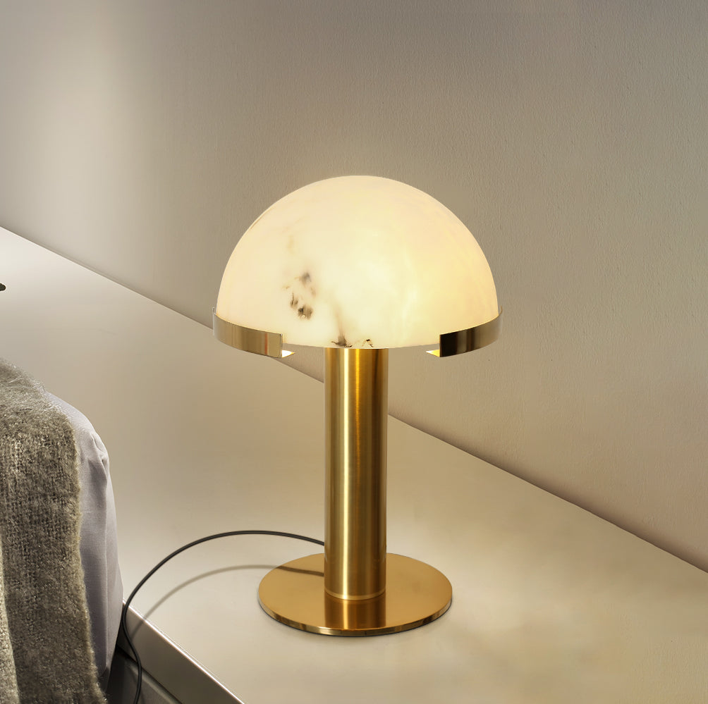 Lit gold Clérachic table lamp on a pale ledge, seen from a low angle with a knitted throw and black cord beside it.
