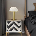 Gold Clérachic table lamp standing tall above a black and white chevron nightstand, with a brass bedpost and dark bedding nearby.