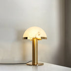 Gold Clérachic table lamp glowing on a light surface in a soft cream room, with the cord trailing across to the right.