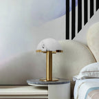 Gold Clérachic table lamp on a round side table with a grey marble top, in front of a curved cream headboard.