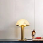 Gold Clérachic table lamp lit on a dark console, with an amber bottle and a glass ball in front of a pale wall.