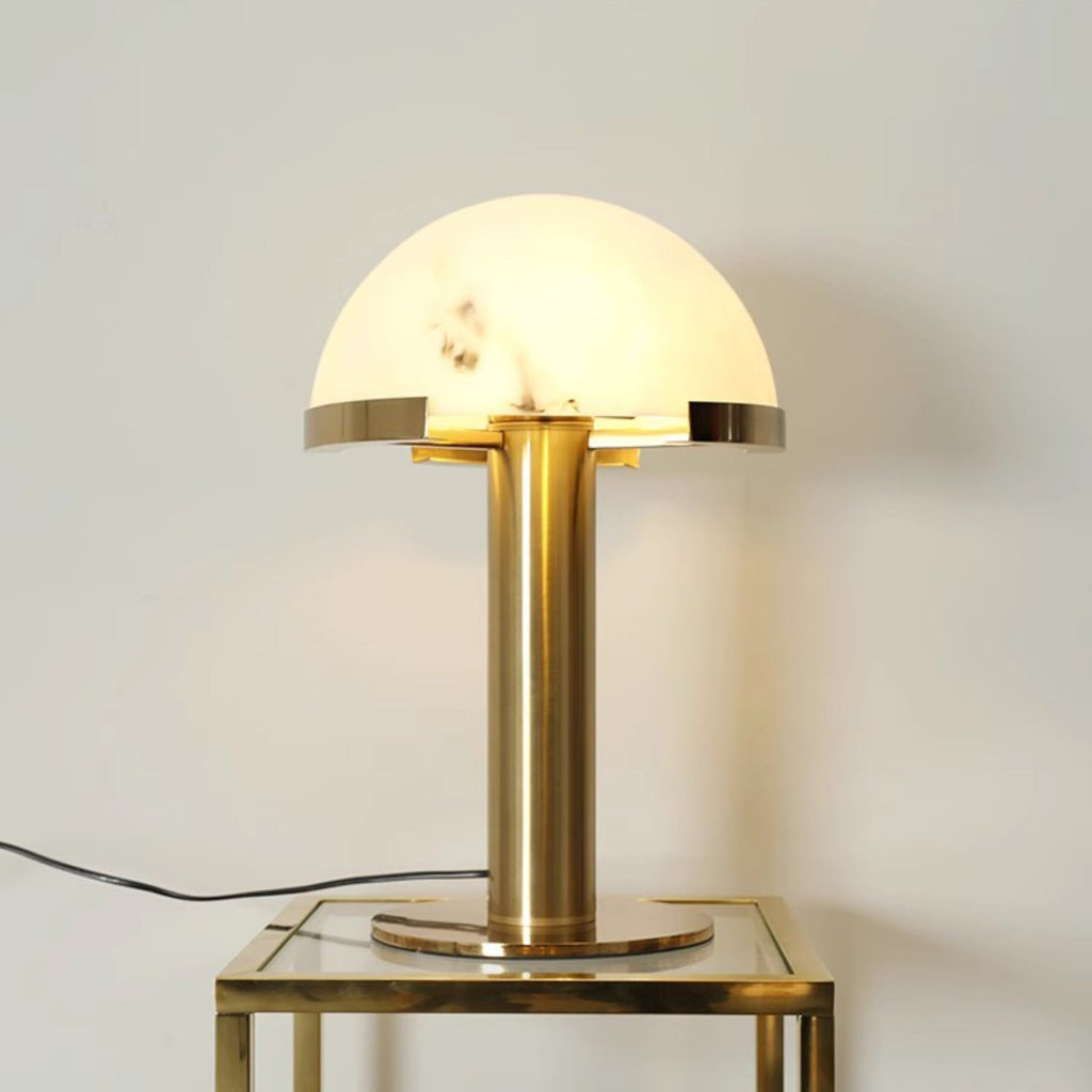 Lit gold Clérachic table lamp on a gold-framed glass side table, glowing against a pale wall with the cord trailing left.