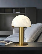 Gold Clérachic table lamp on a dark table in a living room, with a grey sofa and a magazine beside it.