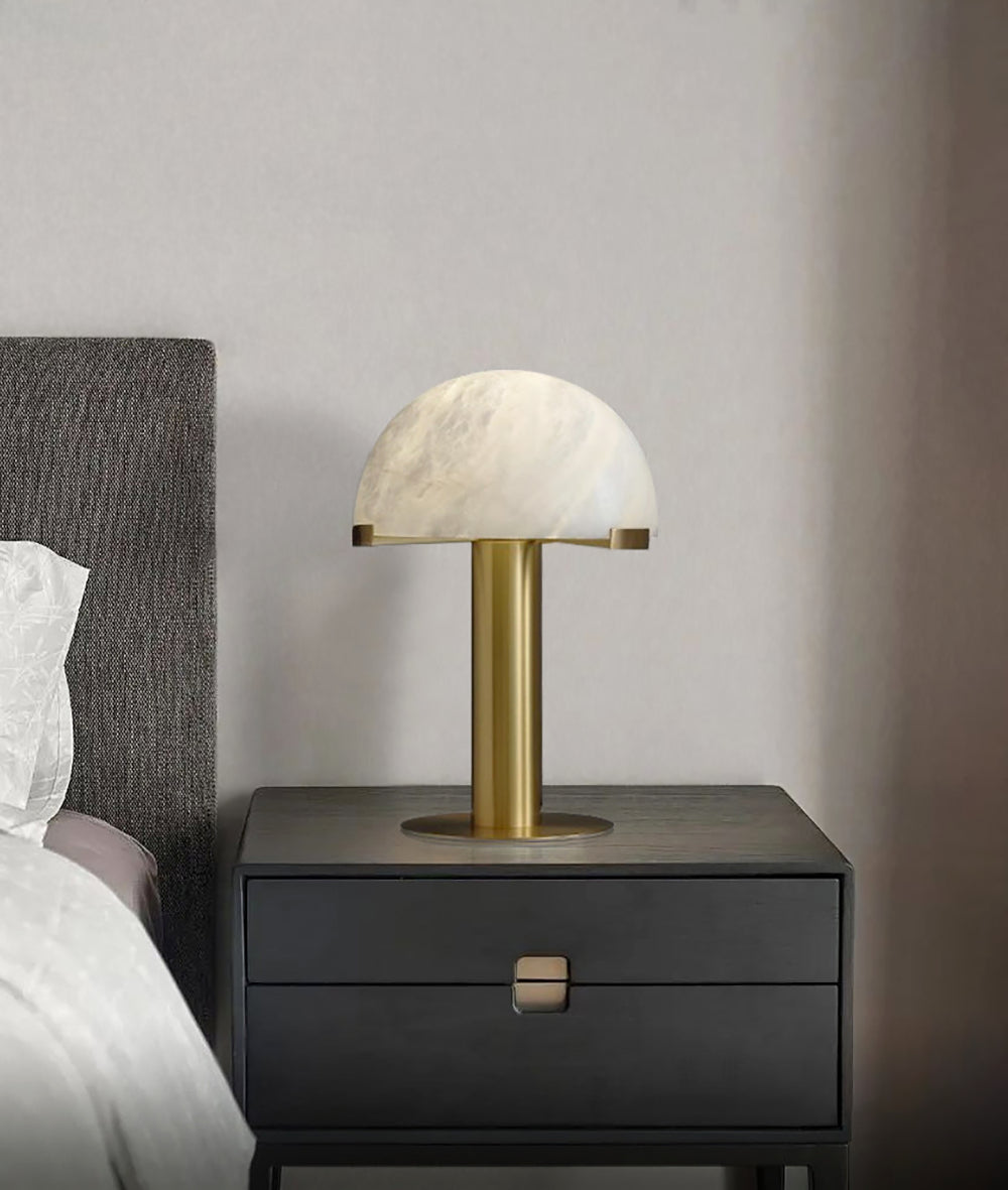 Gold Clérachic table lamp on a black nightstand with a brass pull, in front of a grey headboard and a beige wall.