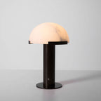Dark bronze-coloured Clérachic table lamp lit on a pale grey backdrop, the dome glowing a warm peach with soft grey veins.