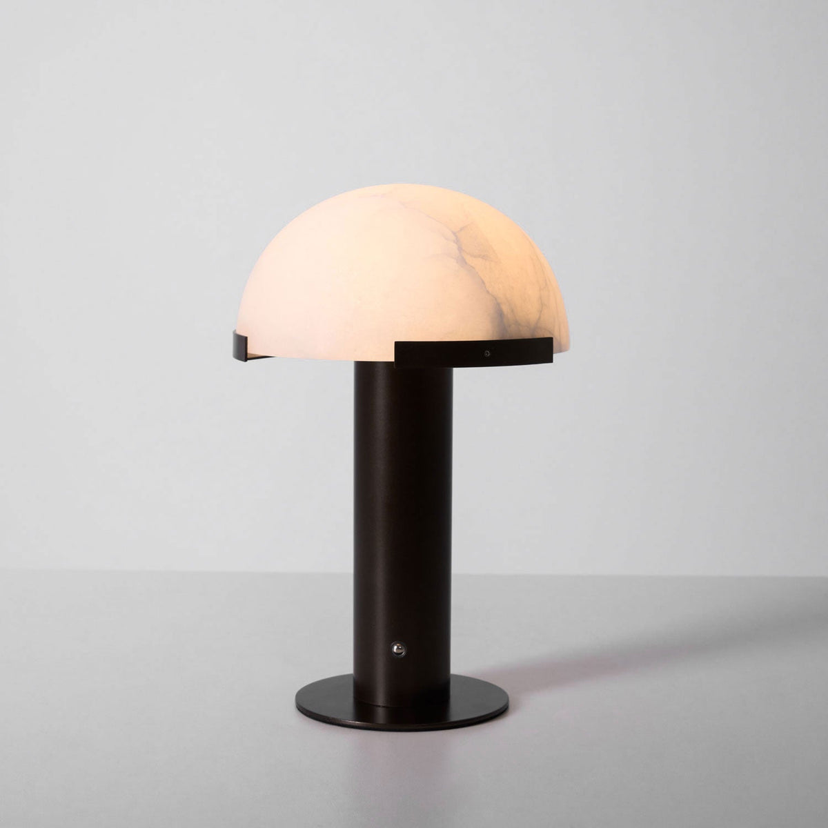 Dark bronze-coloured Clérachic table lamp lit on a pale grey backdrop, the dome glowing a warm peach with soft grey veins.