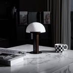 Dark bronze-coloured Clérachic table lamp on a white marble table, with a chequered mug, art books and dark shelves behind.
