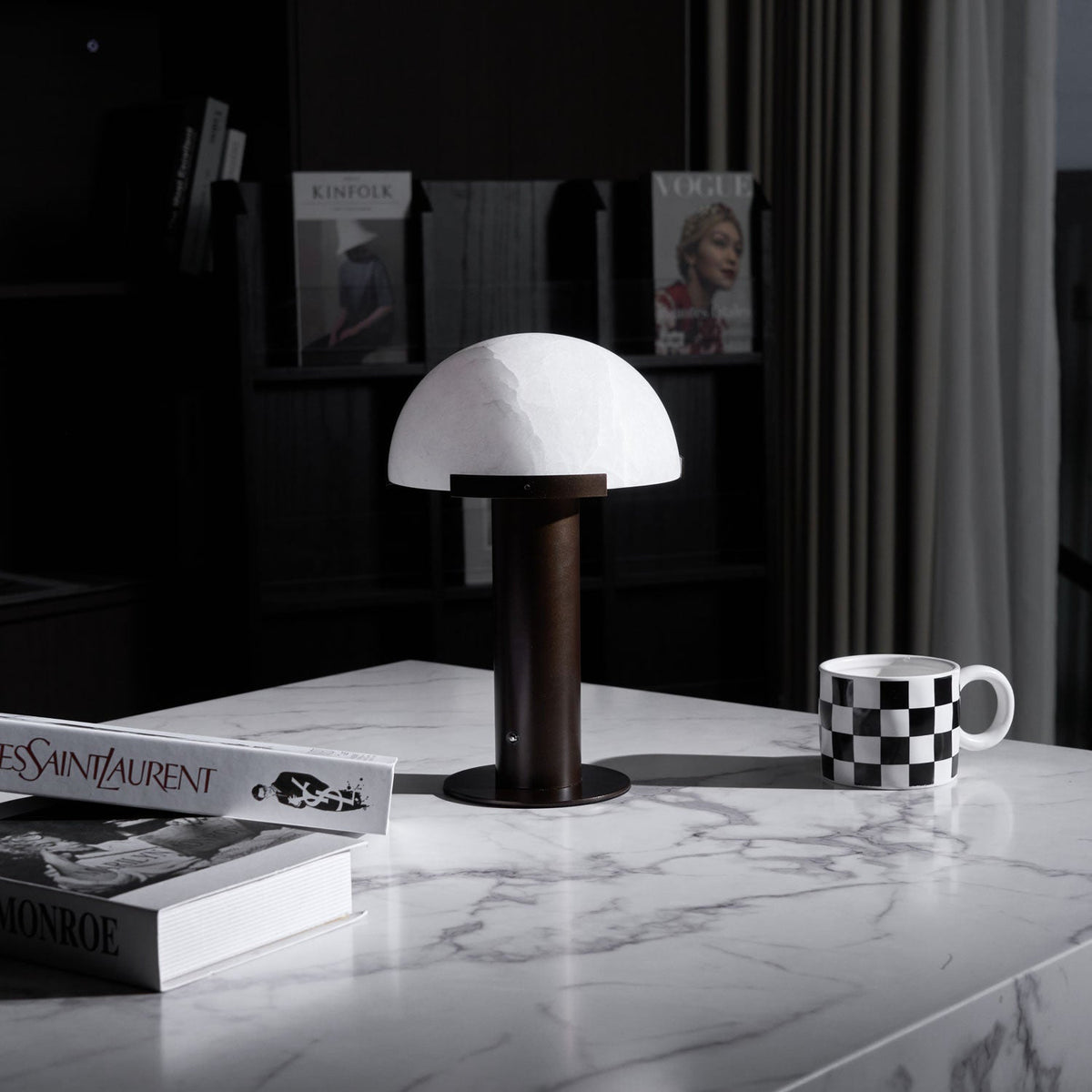 Dark bronze-coloured Clérachic table lamp on a white marble table, with a chequered mug, art books and dark shelves behind.
