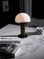Narrow view of the dark-based Clérachic table lamp lit softly on a marble table, with a chequered mug and books to the sides.