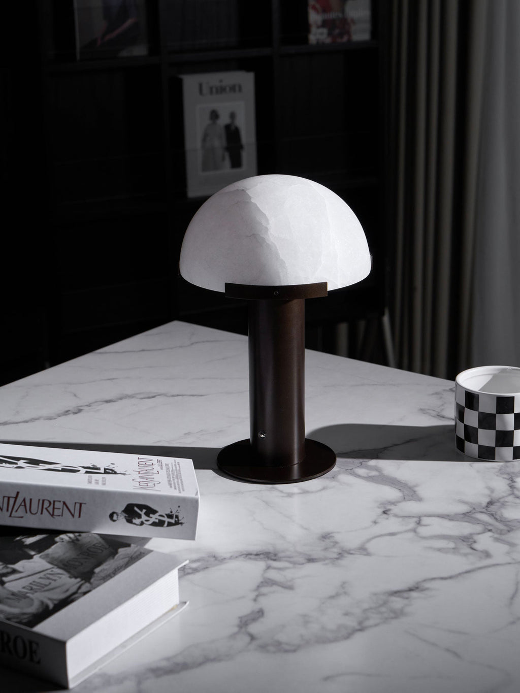 Dark bronze-coloured Clérachic table lamp on a white marble table, seen from a higher angle with books and a mug at the edges.