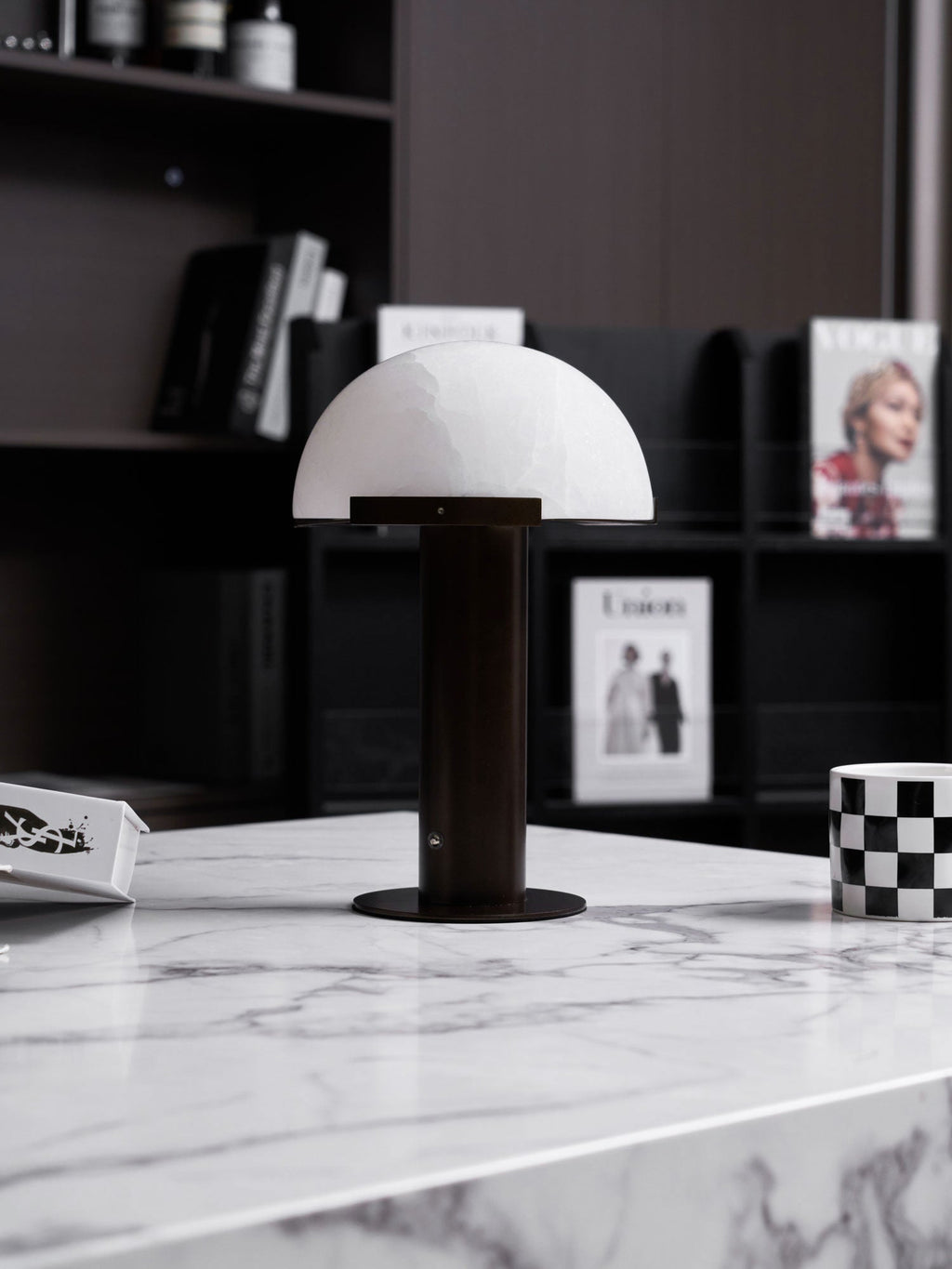 Dark-based Clérachic table lamp with a white dome on a marble table edge, in front of black shelving with magazines.