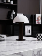 Dark-based Clérachic table lamp with a white dome on a marble table edge, in front of black shelving with magazines.