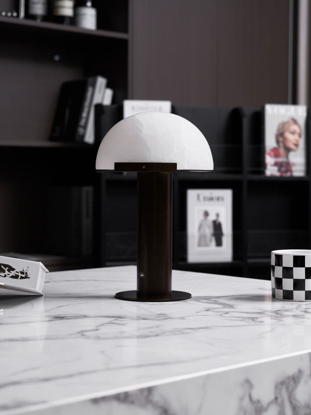 Dark-based Clérachic table lamp with a white dome on a marble table edge, in front of black shelving with magazines.