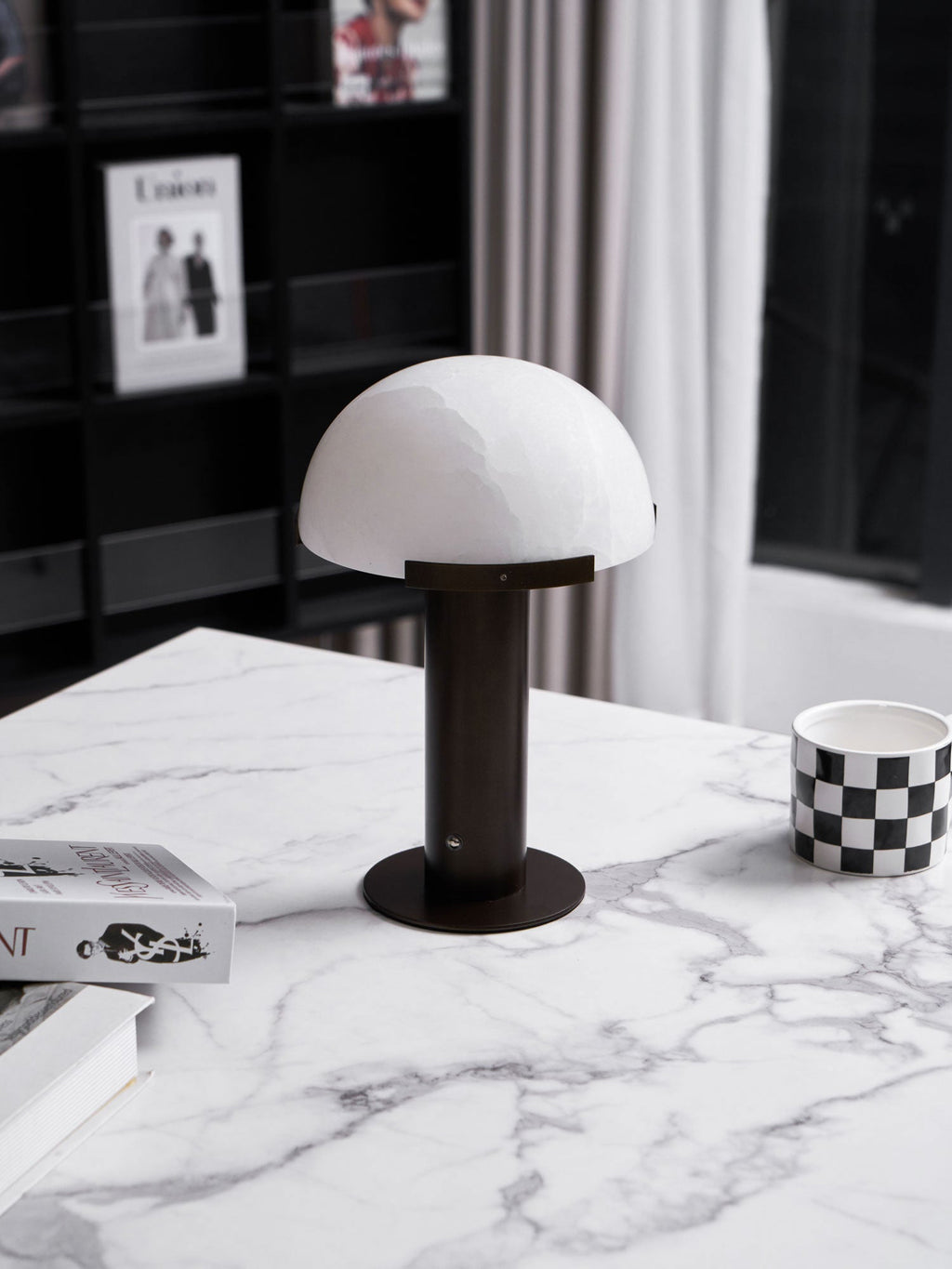 Dark bronze-coloured Clérachic table lamp at a low angle on a marble top, with a chequered mug and a stack of books beside it.