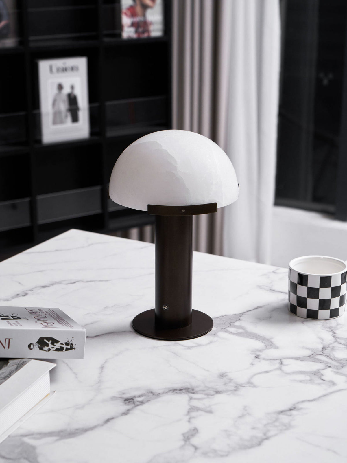 Dark bronze-coloured Clérachic table lamp at a low angle on a marble top, with a chequered mug and a stack of books beside it.