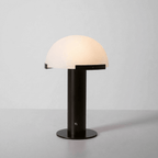 Black-based Clérachic table lamp lit on a pale grey backdrop, its dome glowing a soft cream above a round black base.