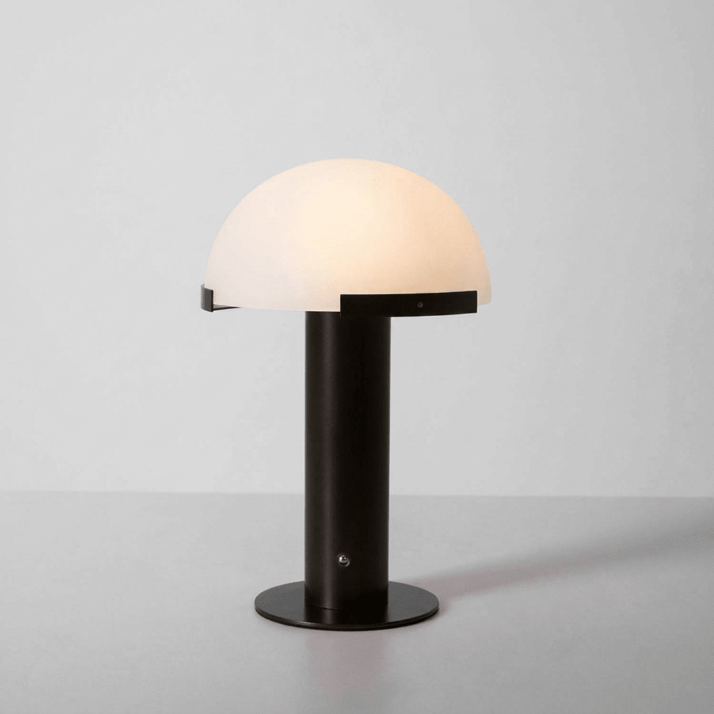 Black-based Clérachic table lamp lit on a pale grey backdrop, its dome glowing a soft cream above a round black base.
