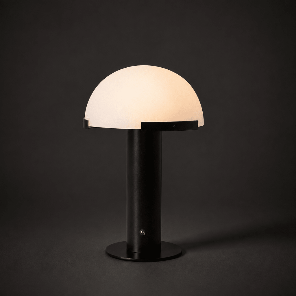 Black Clérachic table lamp lit in a dark setting, with a glowing cream dome and a small round fitting on the stem.