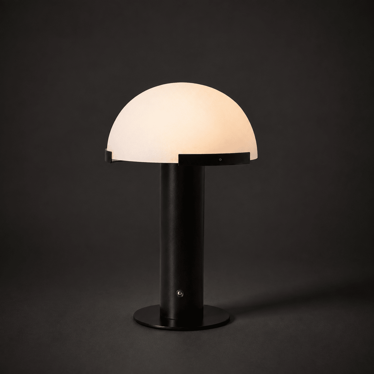 Black Clérachic table lamp lit in a dark setting, with a glowing cream dome and a small round fitting on the stem.