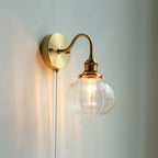 The Clérachic Pull-Chain Brass Wall Sconce lit on a pale grey-blue wall, its clear fluted glass globe showing the filament bulb inside.