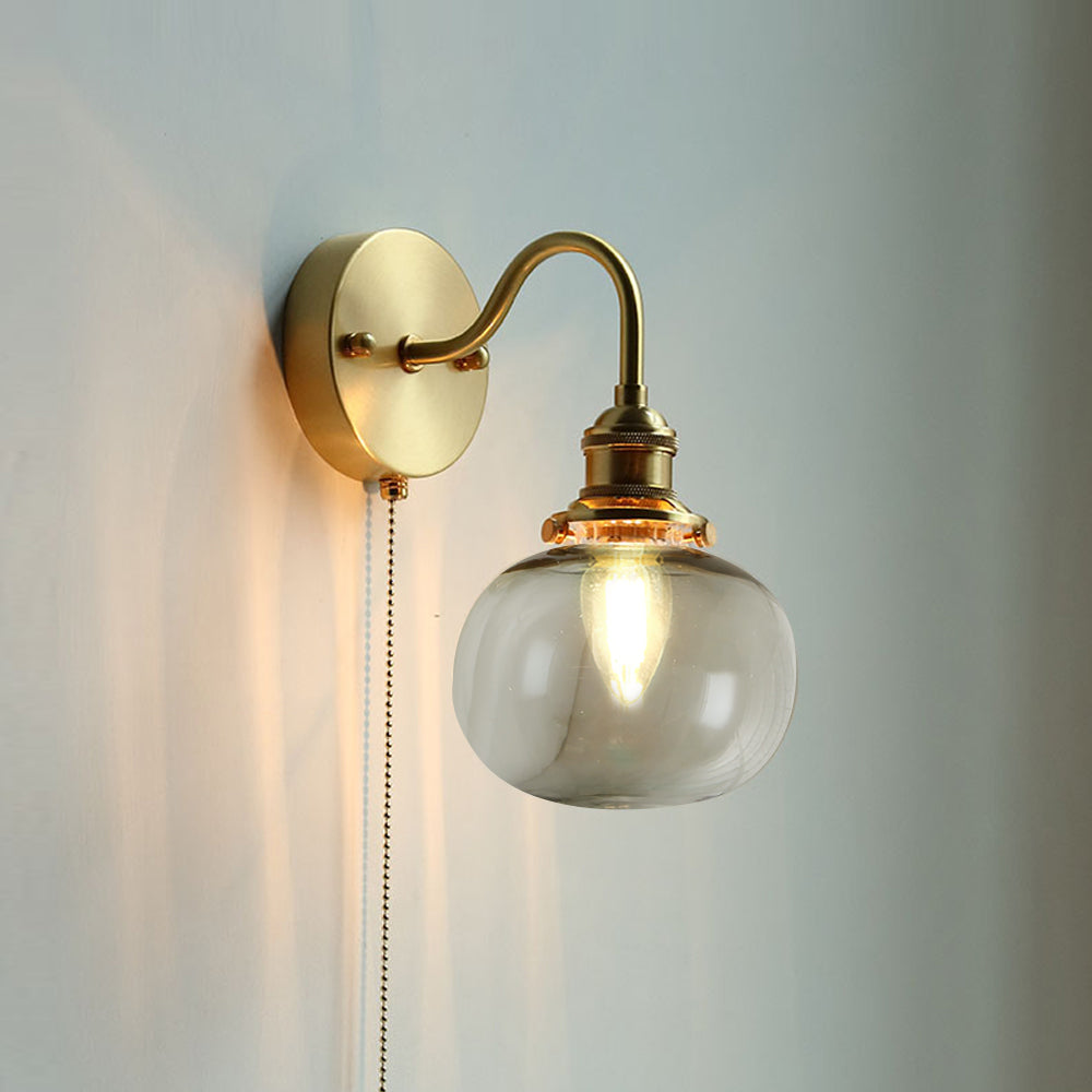 Clérachic Pull-Chain Brass Wall Sconce in smoke gray glass, the smooth squat globe warming the light against a soft grey-blue wall.