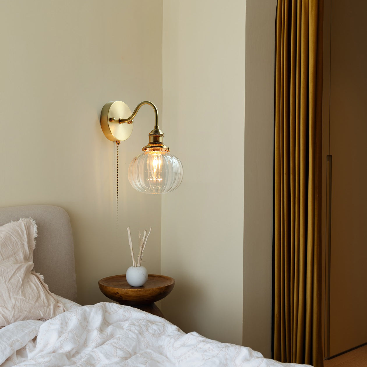 The Clérachic brass wall sconce lit above a bedside table, its clear fluted globe casting a warm pool of light over the bed linen.