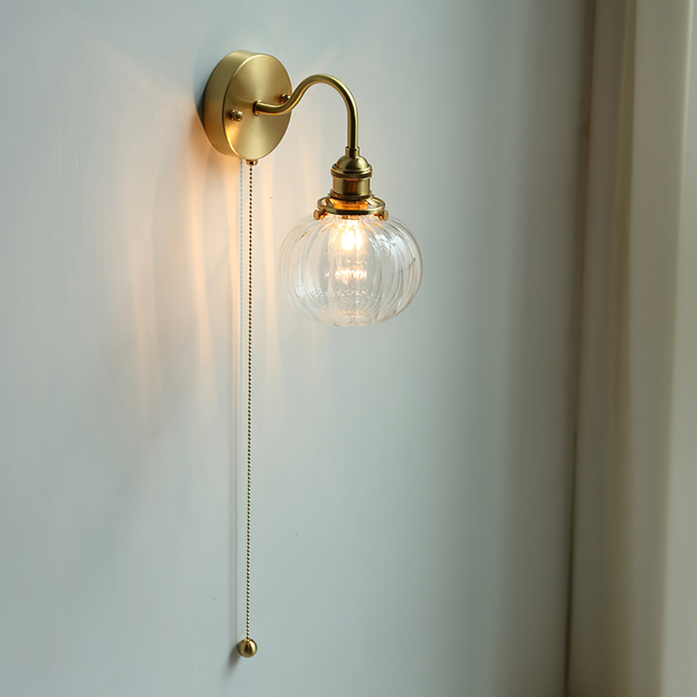 Clérachic brass sconce lit on a pale wall, the bead pull chain ending in a small brass ball you can find in the dark.