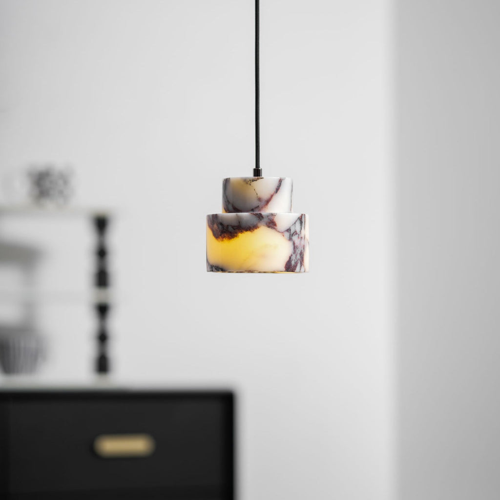 A small marble Clérachic pendant hangs in a soft-focus room, with a black chest of drawers and brass handles below.