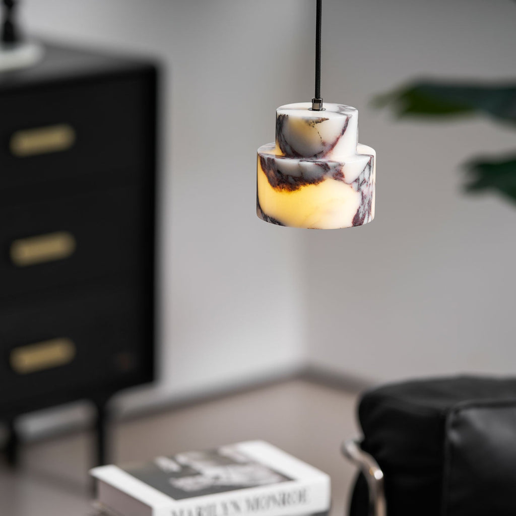 The two stepped marble tiers of this Clérachic pendant glow amber, with a black chest and a stack of books blurred below.