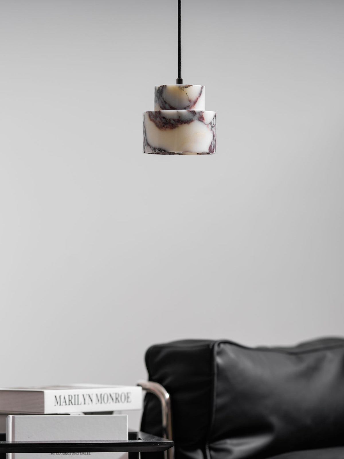 A small, unlit marble Clérachic pendant hangs against a pale wall, above a leather armchair and a book on a side table.