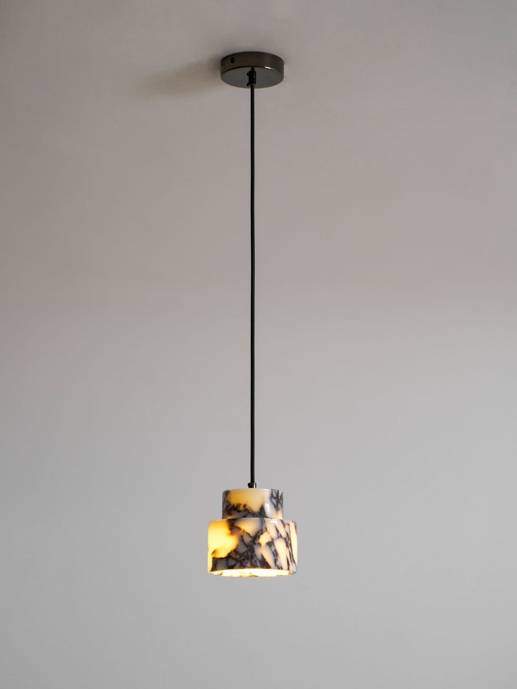 A small marble Clérachic pendant on a black cord and a dark ceiling cup, lit warm against a soft grey wall.