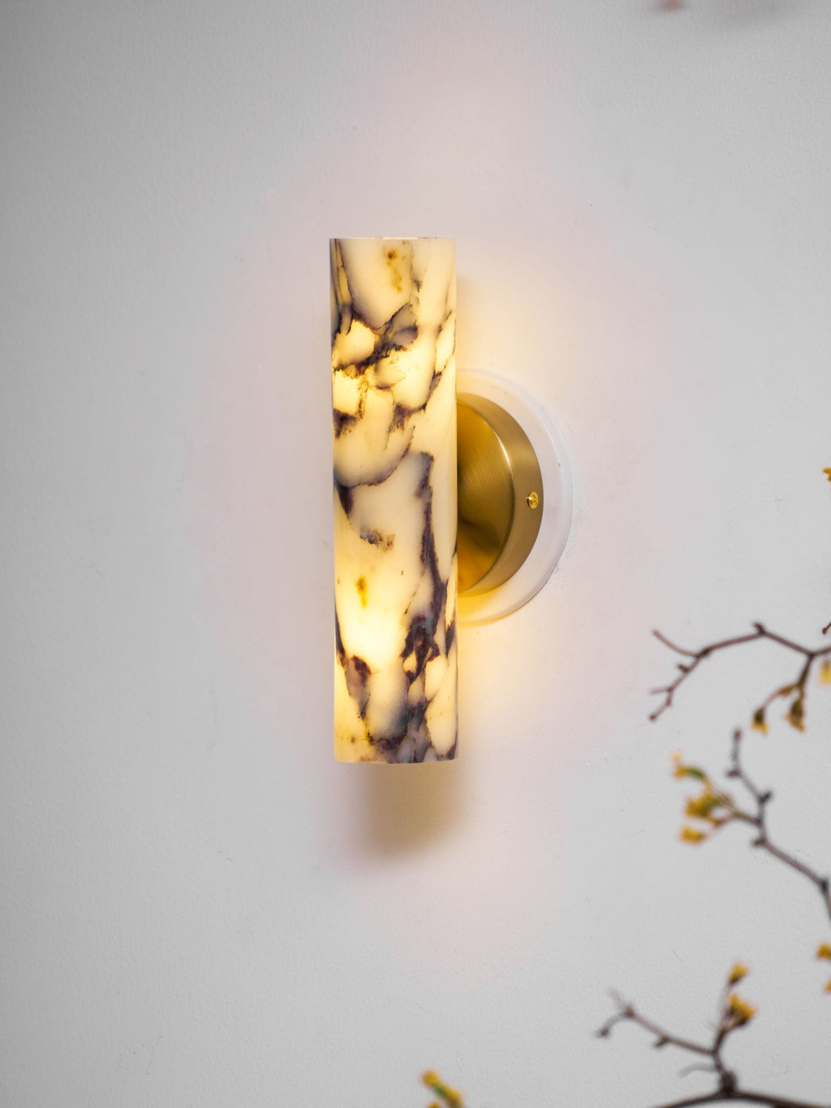 Clérachic wall light switched on beside a bare flowering branch, the purple-veined marble shade glowing on a pale wall.