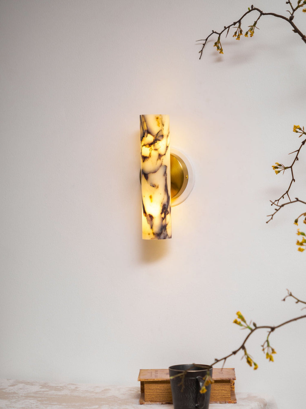 Small in the frame, this lit Clérachic wall light hangs above a shelf with a dark mug on a book, with blossom twigs at the edges.