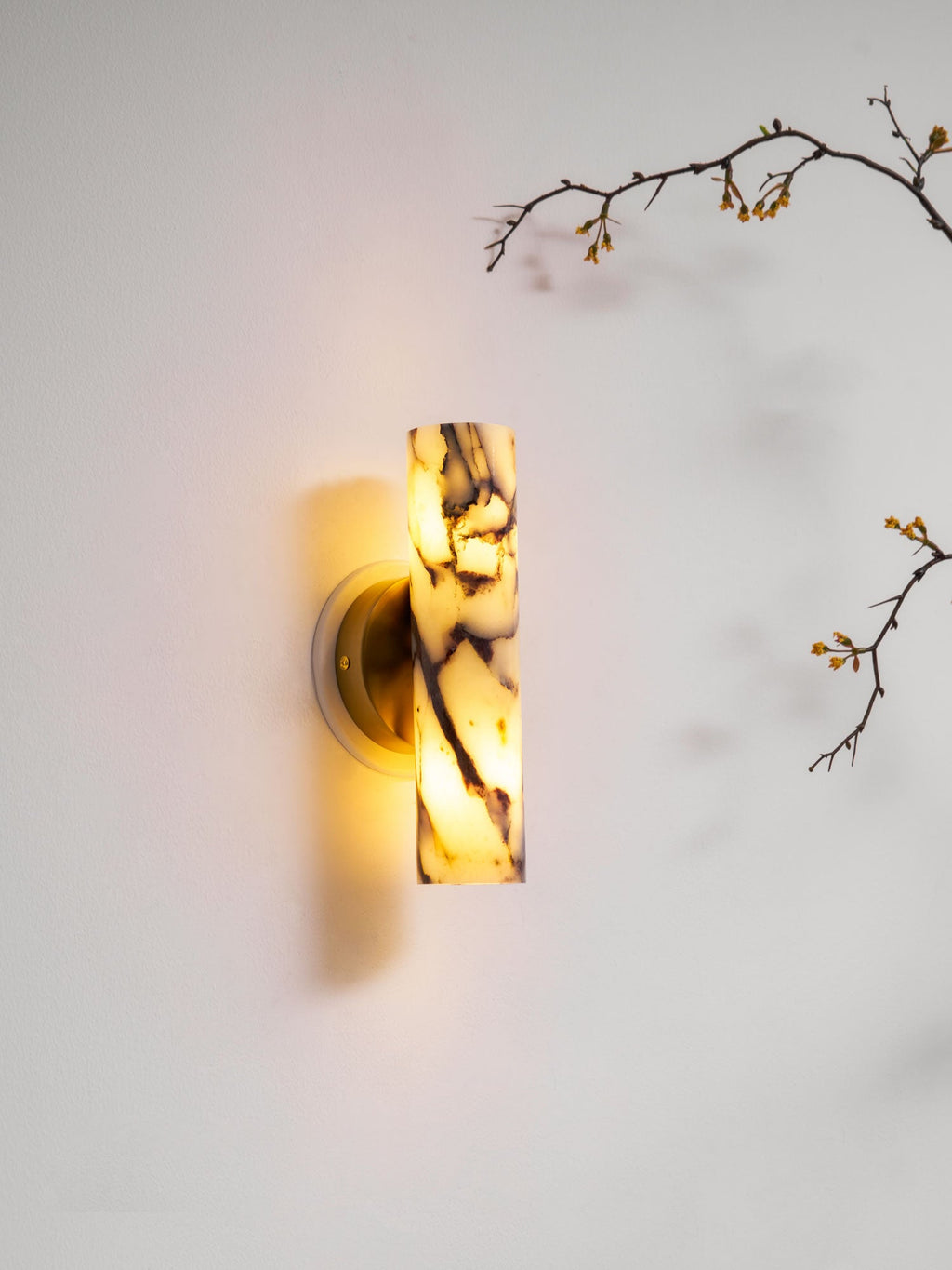 Marble wall light from Clérachic lit and mounted low on a pale wall, with a blossom branch reaching in from the top right.