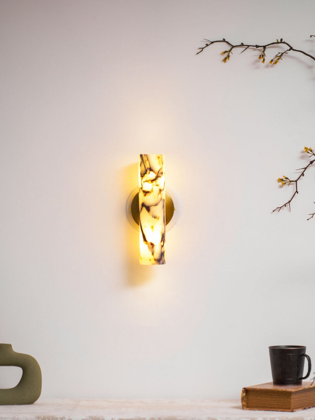 Distant view of a lit Clérachic marble wall light above a shelf with a green sculptural vase and a mug on a book.