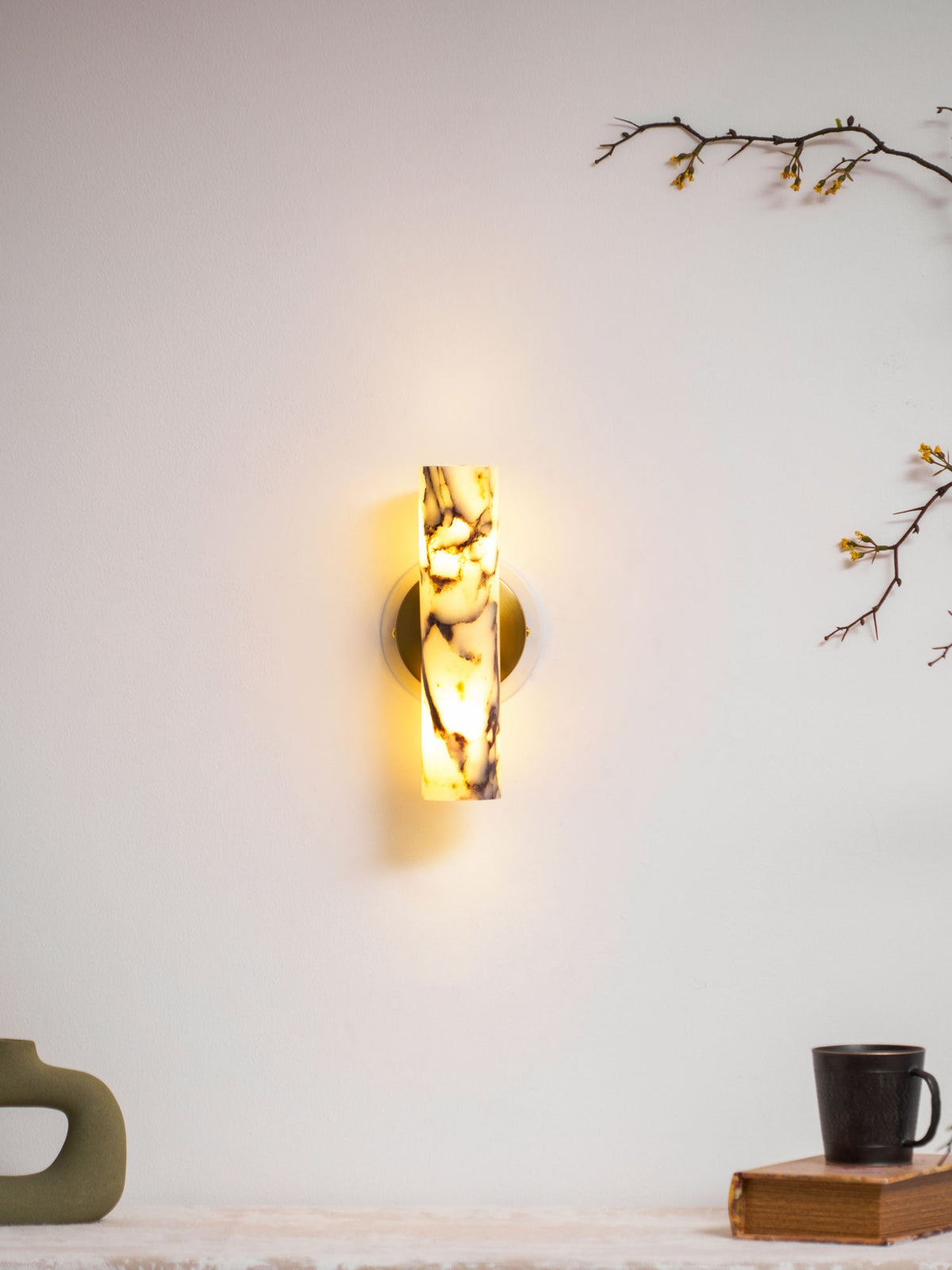 Distant view of a lit Clérachic marble wall light above a shelf with a green sculptural vase and a mug on a book.