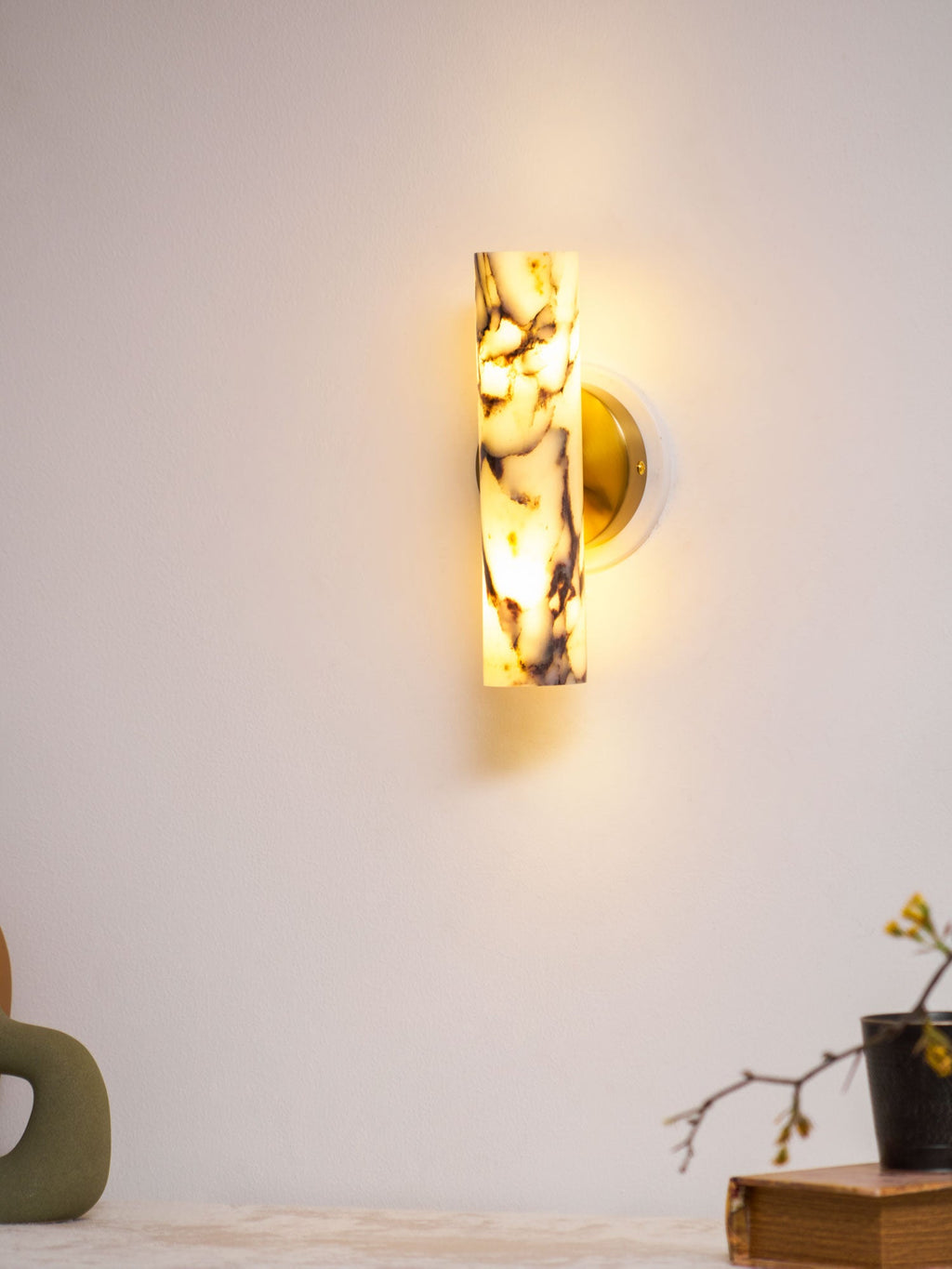 Glowing Clérachic wall light with a purple-veined marble shade, above a green vase and a blossom twig in a small dark pot.