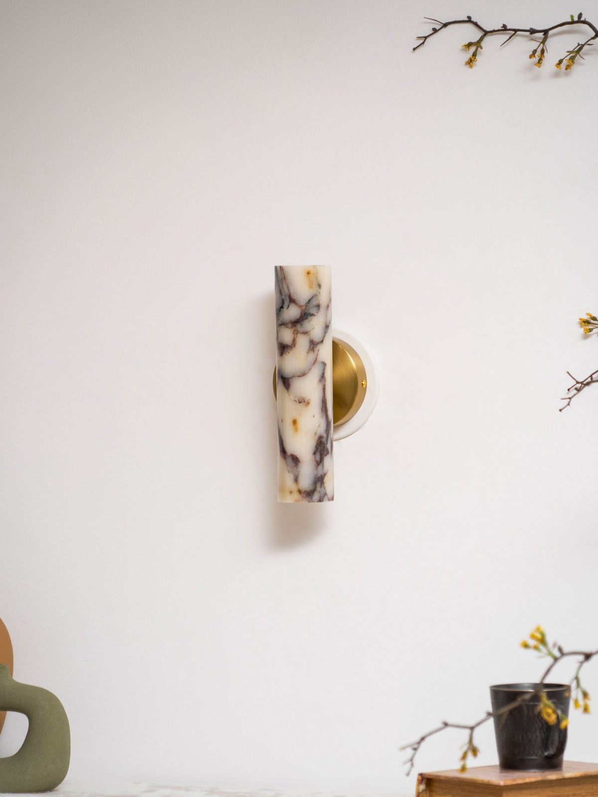Unlit Clérachic wall light with a purple-veined marble shade, on a white wall above a green vase and a mug on a book.