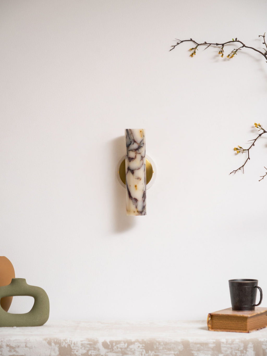 Small unlit Clérachic wall light high on a white wall, with a green vase, a mug on a book and a blossom branch below.