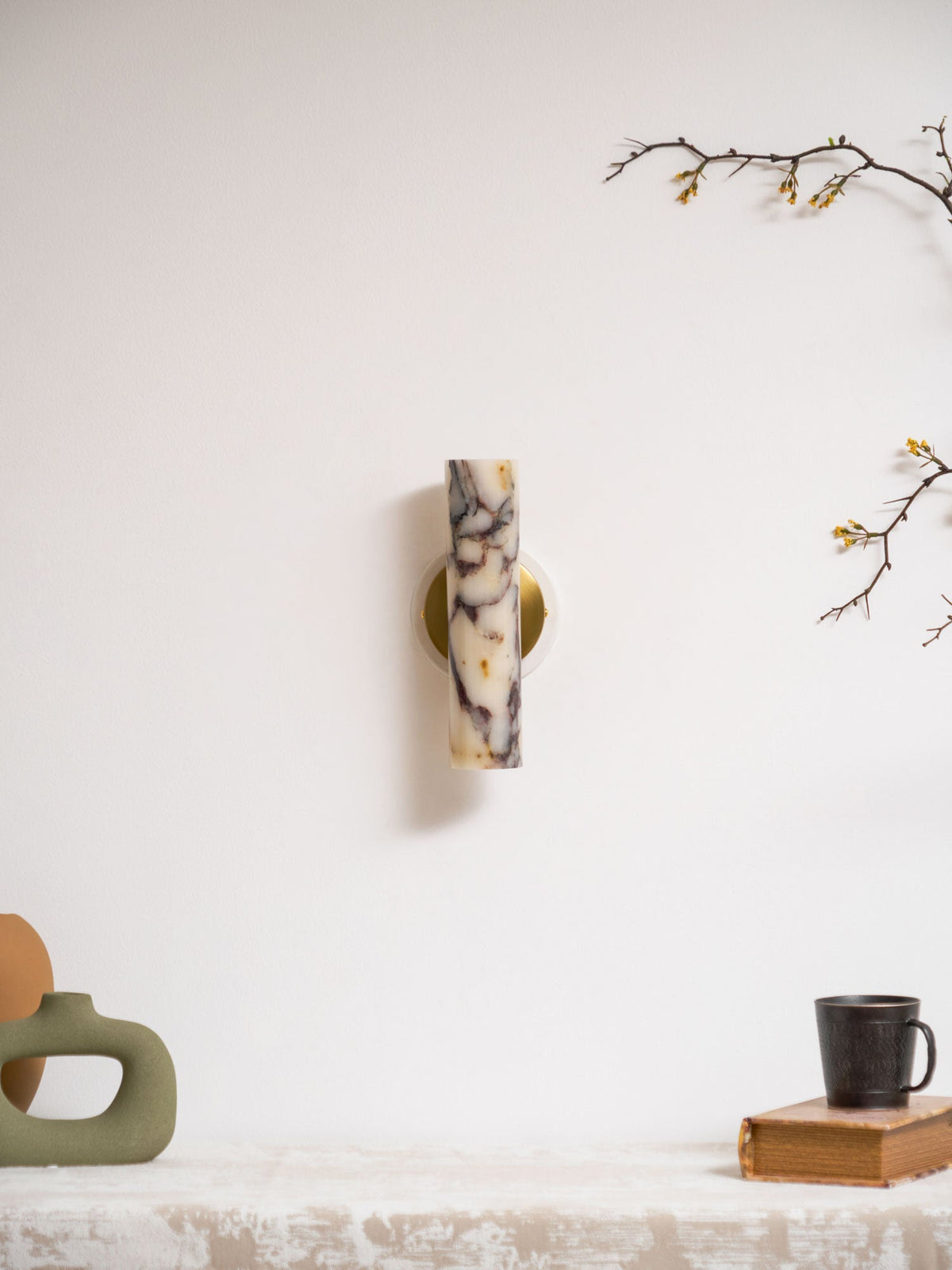 Small unlit Clérachic wall light high on a white wall, with a green vase, a mug on a book and a blossom branch below.